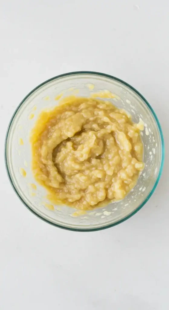 Purée de bananes jaune-beige dans un bol en verre, présentant une texture à la fois lisse et grumeleuse, idéale pour un gâteau pomme banane.