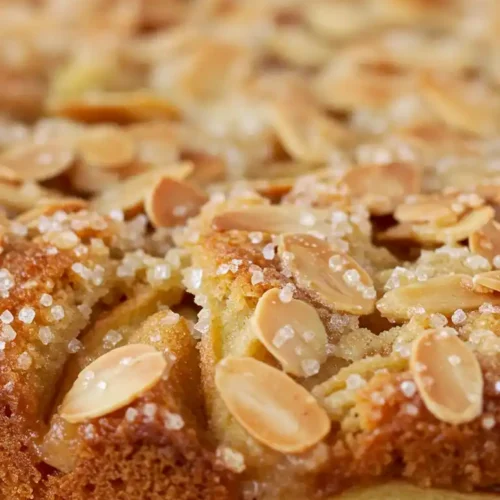 Gros plan d'un délicieux gâteau pomme amandes. On y voit la surface du gâteau, parsemée d'amandes effilées et de cristaux de sucre, avec une croûte dorée et moelleuse sur les côtés. Le gâteau est posé sur une planche à découper en bois. Des feuilles vertes, floues, sont visibles à l'arrière-plan, ajoutant une touche de fraîcheur.