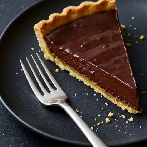 Une part de tarte chocolat, avec sa garniture brillante, est présentée sur une assiette noire, accompagnée d'une fourchette. La tarte est prête à être dégustée.