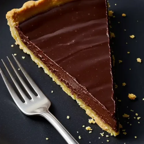 Une délicieuse part de tarte chocolat, avec sa croûte dorée et sa ganache onctueuse, est prête à être savourée sur une assiette noire, accompagnée d'une fourchette.