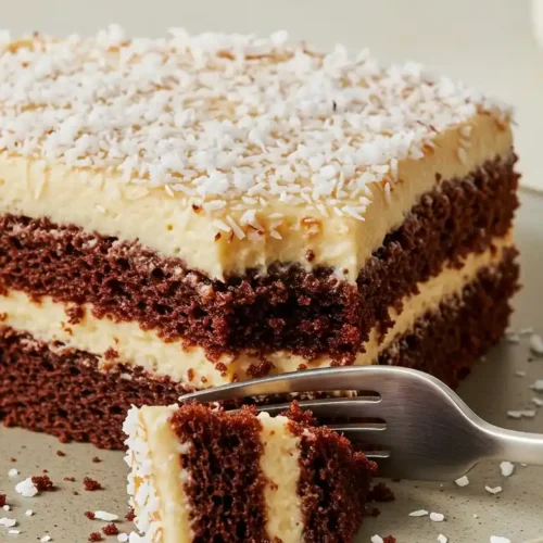 Une tranche de gâteau à la noix de coco et chocolat, présentée sur une assiette en céramique, est coupée à l'aide d'une fourchette. On observe les deux couches de gâteau au chocolat séparées par une couche de crème vanillée, le tout recouvert d'une épaisse couche de glaçage blanc et saupoudré de noix de coco râpée. Des miettes de gâteau et de noix de coco se trouvent éparpillées sur l'assiette.