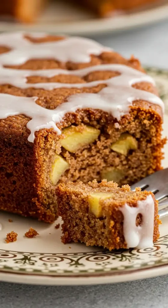 A slice of apple banana cake, visibly moist and studded with pieces of baked apples, placed on a decorative ceramic plate. A silver fork is embedded in the cake. Another slice of apple banana cake is visible in the background