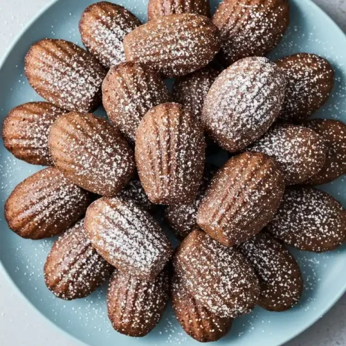 Vue de dessus d'une assiette bleue élégante, généreusement remplie de madeleines au chocolat saupoudrées de sucre glace, avec un fond gris clair.