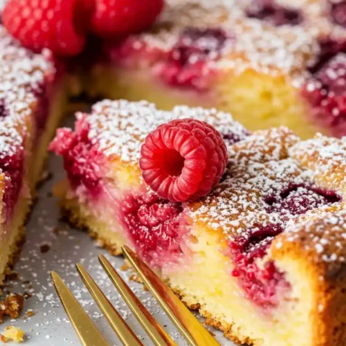 Dégustez une part de Clafoutis Aux Framboises, un dessert français rustique, où les framboises fraîches se marient harmonieusement à une pâte crémeuse, le tout subtilement saupoudré de sucre glace, invitant à une pause gourmande avec une fourchette dorée à portée de main.