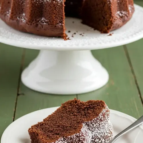 Sur une table verte rustique, on aperçoit un délicieux Gâteau Yaourt Cacao, saupoudré de sucre glace. Une part est présentée sur une assiette blanche avec une fourchette, tandis que le reste du gâteau est élégamment posé sur un présentoir à gâteau en arrière-plan.