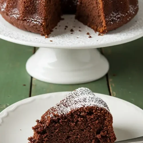 Sur une table en bois verte, une part de Gâteau Yaourt Cacao saupoudrée de sucre glace repose sur une assiette blanche, accompagnée d'une fourchette. En arrière-plan, le reste du gâteau est présenté sur un présentoir à pied, également saupoudré de sucre glace, avec une part manquante.