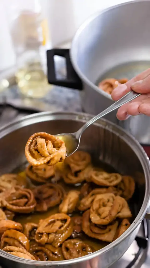 Une chebakia dorée est délicatement extraite d'une casserole remplie d'autres chebakias en train de frire dans de l'huile chaude. Cette étape cruciale confère à la pâtisserie marocaine sa texture croustillante et sa couleur appétissante, avant d'être trempée dans du miel parfumé. On aperçoit également une autre casserole, probablement contenant les prochaines chebakias à frire.