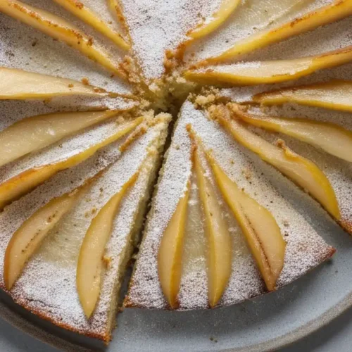 Image en vue plongeante d'un gâteau au yaourt poire, saupoudré de sucre glace et coupé en parts. Les tranches de poires sont visibles sur le dessus du gâteau, créant un motif rayonnant et appétissant. Le gâteau est présenté sur une assiette grise