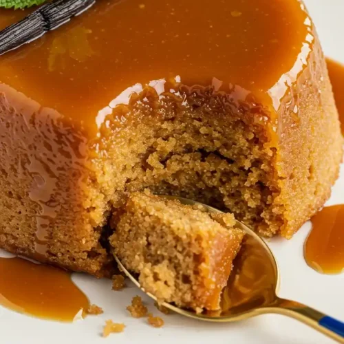 Vue rapprochée d'un délicieux gâteau de semoule au caramel, orné d'une gousse de vanille et de menthe fraîche, avec une part retirée par une cuillère dorée, révélant une texture moelleuse et une sauce caramel dégoulinante.