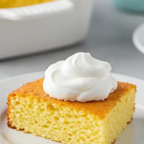 Gros plan sur une part de gâteau yaourt grec, moelleuse et dorée, disposée sur une assiette blanche et garnie d'une généreuse quenelle de crème fouettée. L'arrière-plan flou montre le reste du gâteau dans son plat de cuisson blanc. Une fourchette dorée est partiellement visible en bas de l'image.