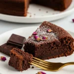 Une part de fondant au chocolat Nestlé, saupoudrée de sucre glace et agrémentée de pétales de rose, est présentée sur une assiette blanche avec une fourchette dorée. Un morceau du gâteau est empalé sur la fourchette, prêt à être dégusté. Des carrés de chocolat sont disposés à côté. En arrière-plan, un fondant au chocolat Nestlé entier trône sur une autre assiette.