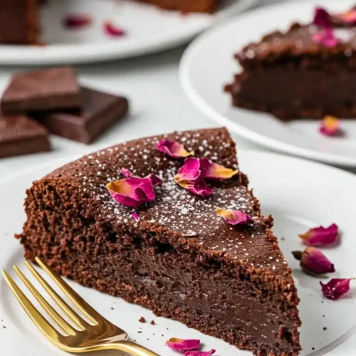 Une part appétissante de fondant au chocolat Nestlé sur une assiette blanche, décorée de sucre glace et de délicats pétales de rose. Une fourchette dorée est placée à côté. En arrière-plan, un fondant au chocolat Nestlé entier et une autre part sont également présentés.