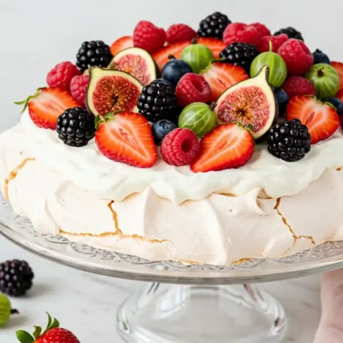 Une somptueuse pavlova recette, garnie d'une généreuse crème fouettée et d'un assortiment coloré de fruits frais : fraises, framboises, mûres, figues, myrtilles et groseilles à maquereau. Le dessert est présenté sur un élégant présentoir en verre, avec quelques fruits éparpillés autour, invitant à la dégustation